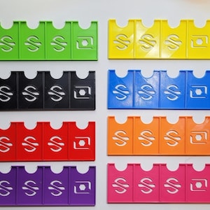 Puede incluir: Conjunto de diez fichas de plástico de colores con un signo de dólar y un diseño hexagonal. Cada ficha está dividida en cinco secciones.