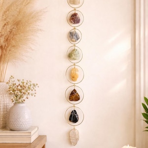 May include: A vertical wall hanging featuring eight raw, multi-colored stones individually wrapped in thin gold-toned wire. Each stone is suspended within a circular gold-toned metal ring, arranged in a single column ending with a dangling stone.