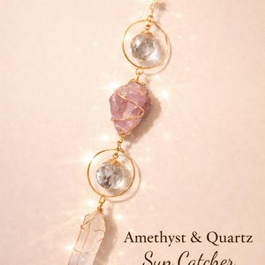 May include: A vertical sun catcher featuring gold-toned wire wrapping, two clear faceted crystal spheres, a raw purple amethyst stone, and a clear quartz point. A small gold sun charm is attached at the top. Text reads: Amethyst & Quartz Sun Catcher.