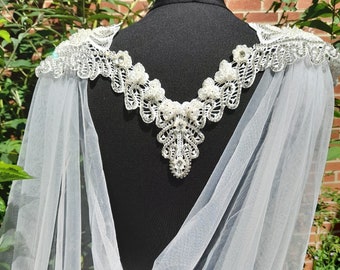 May include: A white tulle bridal cape with a beaded lace neckline. The lace is embellished with pearls and rhinestones.