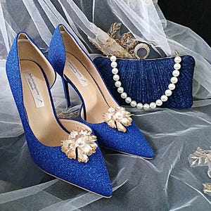 May include: A pair of sparkling blue high heels with gold floral embellishments and a matching clutch with a pearl strap. The shoes have pointed toes and stiletto heels. The clutch is a deep blue color and has a pearl strap.