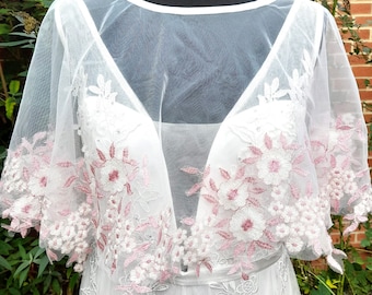 May include: White lace bridal cape with pink floral embroidery. The cape has a sheer, lightweight fabric and a scalloped edge.