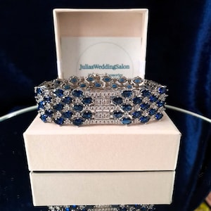 May include: A silver-toned bracelet with multiple rows of blue oval-shaped gemstones and clear accent stones. The bracelet is displayed on a white box with the text "JuliasWeddingSalon" visible. The background is a dark blue.