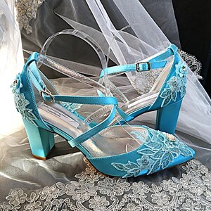 Pode incluir: Um par de sapatos de noiva de cetim turquesa com detalhes de renda floral. Os saltos s&atilde;o em bloco e as tiras s&atilde;o transparentes com detalhes prateados. Os sapatos t&ecirc;m bico fino e um pequeno fecho de fivela. O texto "Julian's Wedding Salon Handmade" &eacute; vis&iacute;vel.