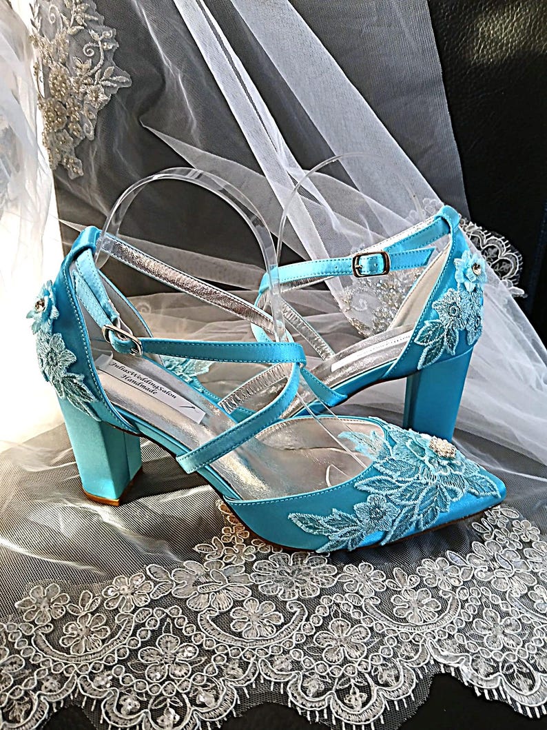 Pode incluir: Um par de sapatos de noiva de cetim turquesa com detalhes de renda floral. Os saltos s&atilde;o em bloco e as tiras s&atilde;o transparentes com detalhes prateados. Os sapatos t&ecirc;m bico fino e um pequeno fecho de fivela. O texto "Julian's Wedding Salon Handmade" &eacute; vis&iacute;vel.