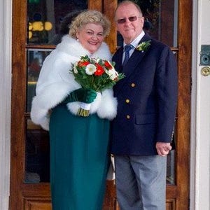 Op de afbeelding: Een stel poseert voor een trouwfoto voor een dubbele houten deur. De vrouw draagt een witte bontjas over een tealgroene jurk en witte schoenen. De man draagt een donkerblauw pak met een wit overhemd en een lichtblauwe das. Ze houden een boeket witte en rode bloemen vast.