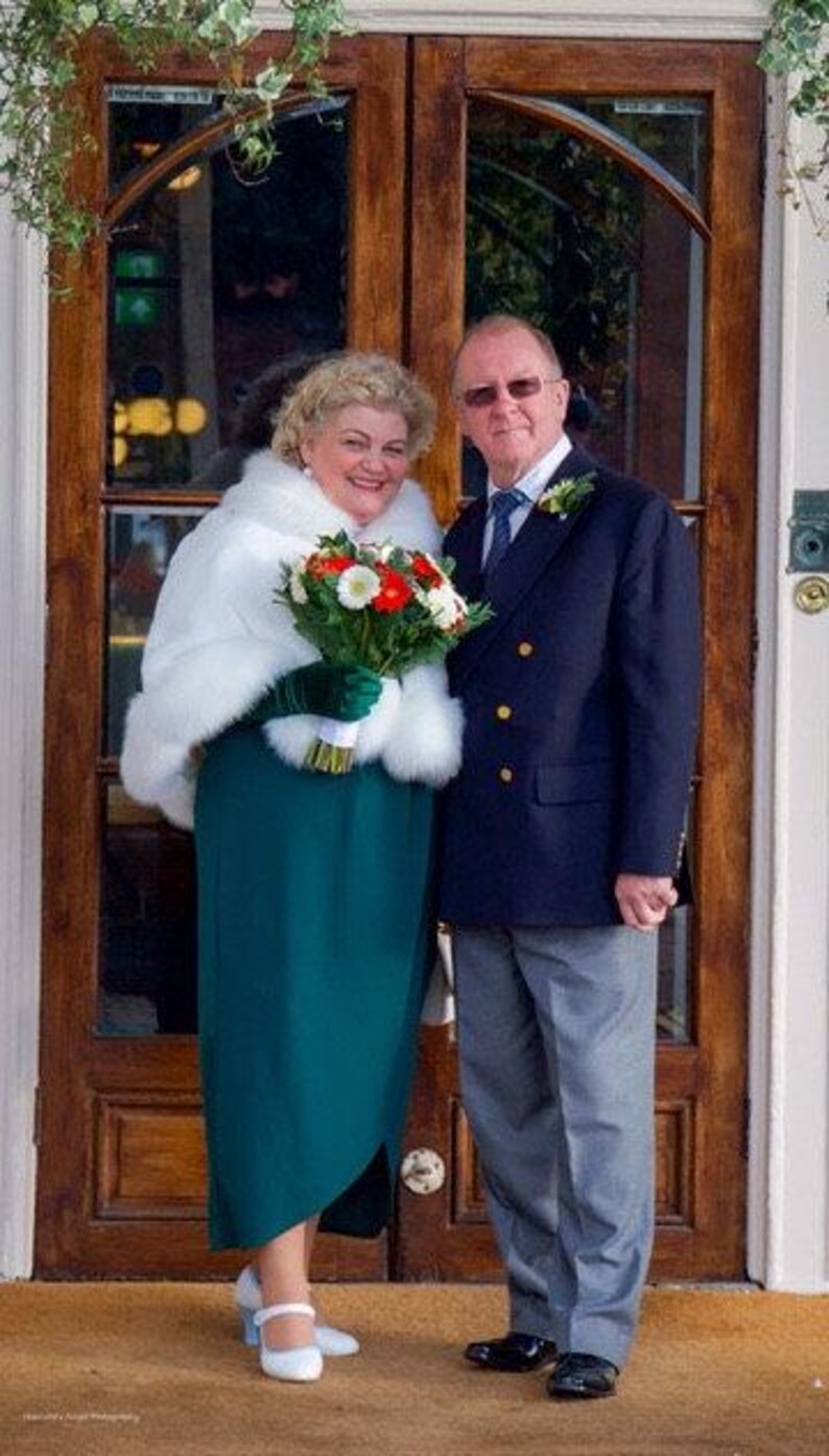 Op de afbeelding: Een stel poseert voor een trouwfoto voor een dubbele houten deur. De vrouw draagt een witte bontjas over een tealgroene jurk en witte schoenen. De man draagt een donkerblauw pak met een wit overhemd en een lichtblauwe das. Ze houden een boeket witte en rode bloemen vast.
