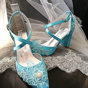 Pode incluir: Um par de sapatos de noiva azul-turquesa com bico fino e salto bloco. Os sapatos apresentam aplica&ccedil;&otilde;es de renda floral e tiras cruzadas. Um v&eacute;u branco com acabamento em renda est&aacute; ao fundo.