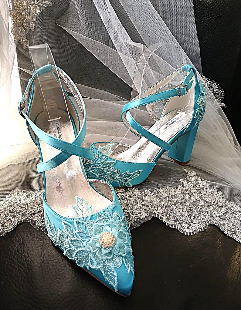 Pode incluir: Um par de sapatos de noiva azul-turquesa com bico fino e salto bloco. Os sapatos apresentam aplica&ccedil;&otilde;es de renda floral e tiras cruzadas. Um v&eacute;u branco com acabamento em renda est&aacute; ao fundo.