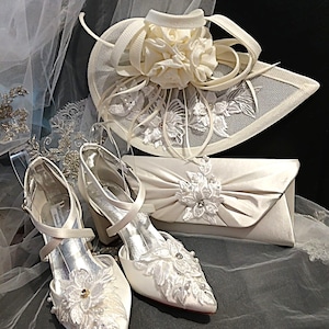 Può includere: Completo da sposa avorio con un paio di scarpe col tacco con dettagli in pizzo floreale, una pochette abbinata e un cappello decorativo. Le scarpe hanno cinturini alla caviglia e la pochette ha un abbellimento floreale.