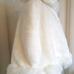 Op de afbeelding: Witte bruidscape van imitatiebont met een brede kraag en een pluche rand. De cape is gemaakt van zacht, pluche materiaal en is perfect om een ​​toets van elegantie toe te voegen aan elke bruidsoutfit.