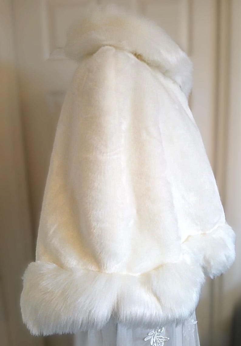 Op de afbeelding: Witte bruidscape van imitatiebont met een brede kraag en een pluche rand. De cape is gemaakt van zacht, pluche materiaal en is perfect om een ​​toets van elegantie toe te voegen aan elke bruidsoutfit.