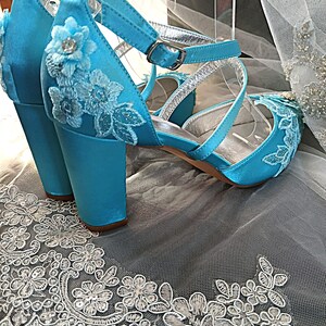 Pode incluir: Um par de sand&aacute;lias de salto alto de cetim turquesa com aplica&ccedil;&otilde;es de renda floral. Os sapatos de bico aberto t&ecirc;m tiras no tornozelo com fivelas prateadas e um salto bloco. Os sapatos s&atilde;o exibidos em tecido de renda branca.
