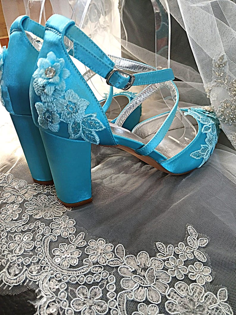Pode incluir: Um par de sand&aacute;lias de salto alto de cetim turquesa com aplica&ccedil;&otilde;es de renda floral. Os sapatos de bico aberto t&ecirc;m tiras no tornozelo com fivelas prateadas e um salto bloco. Os sapatos s&atilde;o exibidos em tecido de renda branca.