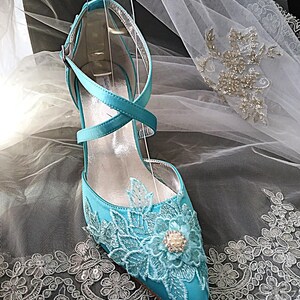 Pode incluir: Um sapato de noiva de cetim turquesa com bico fino e um delicado design floral de renda. O sapato apresenta um fecho de tornozelo com tiras cruzadas e um salto pequeno. Um adorno semelhante a uma p&eacute;rola est&aacute; no centro do design floral. O sapato &eacute; exibido em um suporte transparente.