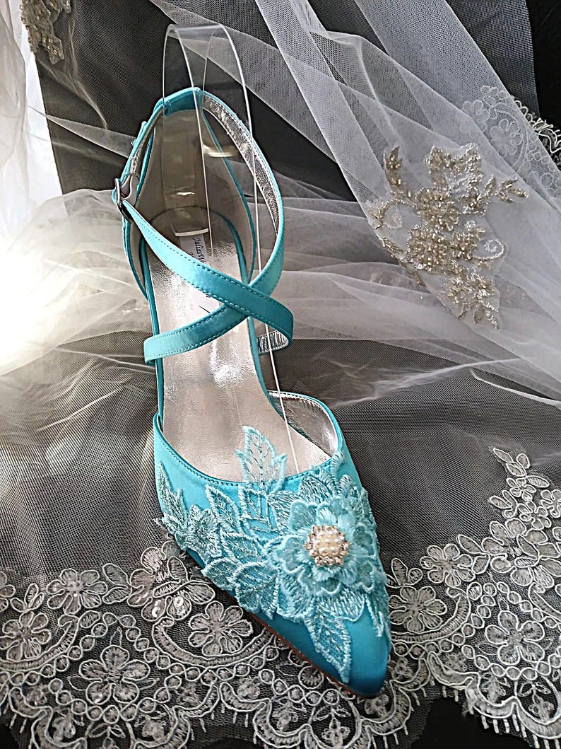 Pode incluir: Um sapato de noiva de cetim turquesa com bico fino e um delicado design floral de renda. O sapato apresenta um fecho de tornozelo com tiras cruzadas e um salto pequeno. Um adorno semelhante a uma p&eacute;rola est&aacute; no centro do design floral. O sapato &eacute; exibido em um suporte transparente.