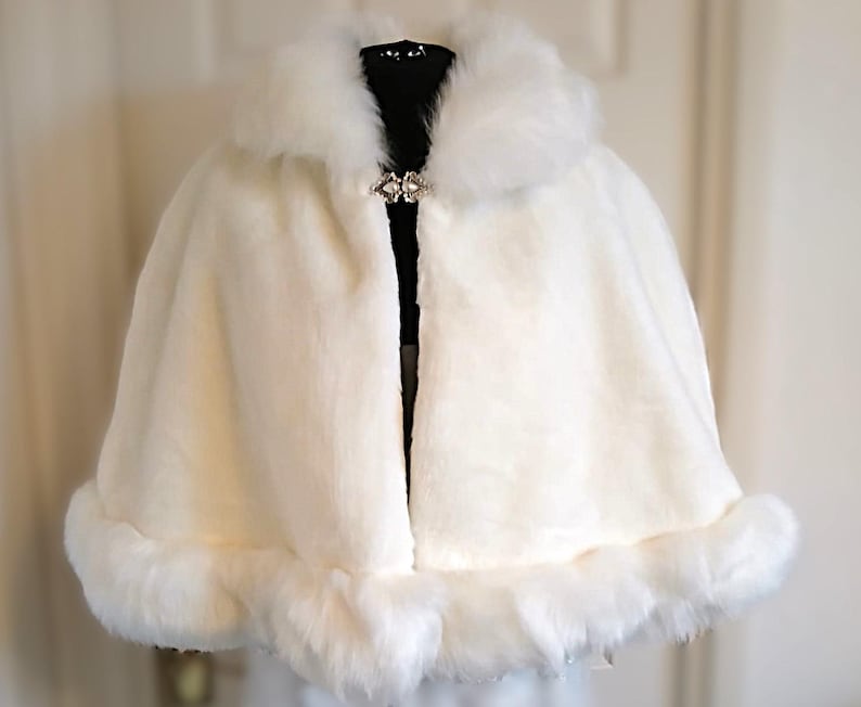 Op de afbeelding: Witte nep bont cape met een grote, pluche kraag en een decoratieve broche. De cape heeft een afgeronde zoom en is afgewerkt met een band van nep bont.