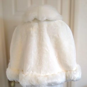 Op de afbeelding: Een witte nep bontjas met een pluche kraag en rand. De jas is gemaakt van zacht, pluche stof en heeft een eenvoudig, elegant ontwerp.