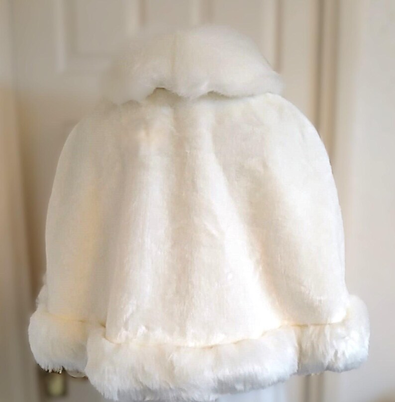 Op de afbeelding: Een witte nep bontjas met een pluche kraag en rand. De jas is gemaakt van zacht, pluche stof en heeft een eenvoudig, elegant ontwerp.