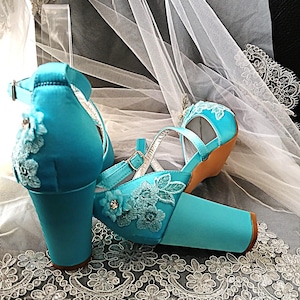 Pode incluir: Um par de sapatos de noiva de cetim turquesa com bordados florais e tiras no tornozelo. Os saltos bloco s&atilde;o adornados com detalhes delicados de renda. Os sapatos s&atilde;o exibidos em um tecido de renda branco.