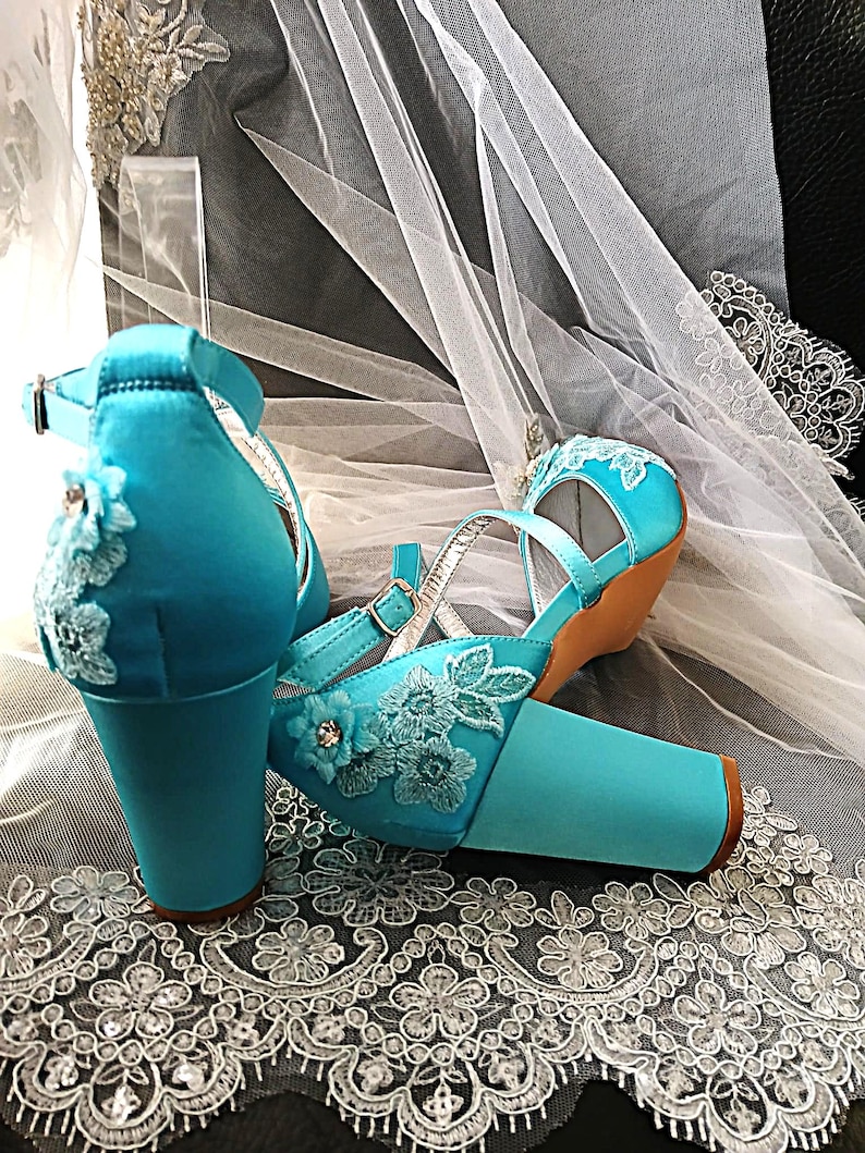 Pode incluir: Um par de sapatos de noiva de cetim turquesa com bordados florais e tiras no tornozelo. Os saltos bloco s&atilde;o adornados com detalhes delicados de renda. Os sapatos s&atilde;o exibidos em um tecido de renda branco.