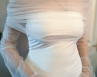 May include: White tulle bridal shrug with long sleeves. The shrug is draped over the shoulders and has a loose, flowing fit.