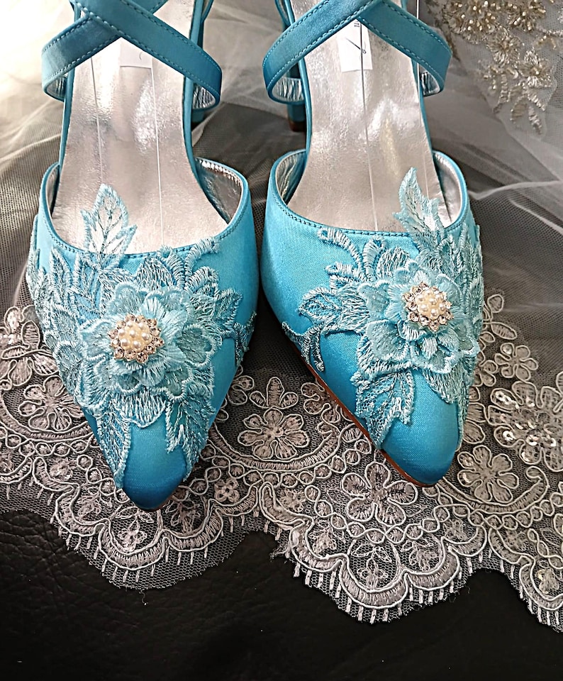 Pode incluir: Um par de sapatos de noiva de cetim azul-turquesa com bordados florais e detalhes de p&eacute;rolas. Os saltos abertos apresentam detalhes delicados em renda e tiras cruzadas. Sapatos para uma ocasi&atilde;o especial.