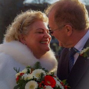 Op de afbeelding: Een bruid en bruidegom die tijdens hun huwelijksceremonie naar elkaar lachen. De bruid draagt een witte bontjas en houdt een boeket met witte en rode bloemen vast. De bruidegom draagt een donker pak en een wit overhemd met een gepatenteerde stropdas.