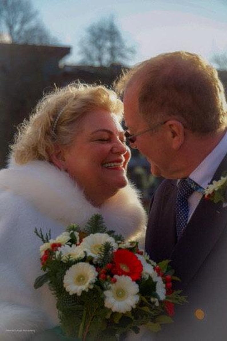 Op de afbeelding: Een bruid en bruidegom die tijdens hun huwelijksceremonie naar elkaar lachen. De bruid draagt een witte bontjas en houdt een boeket met witte en rode bloemen vast. De bruidegom draagt een donker pak en een wit overhemd met een gepatenteerde stropdas.