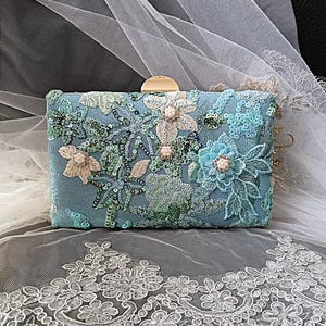 Pode incluir: Uma clutch retangular azul claro com bordados florais e lantejoulas. A clutch apresenta um fecho dourado e &eacute; adornada com flores bordadas em v&aacute;rios tons de azul, verde e bege.
