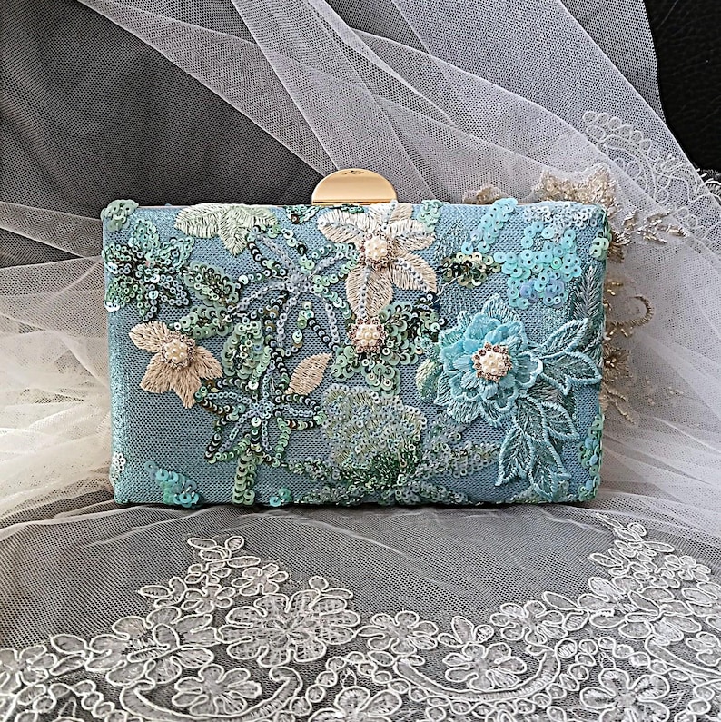 Pode incluir: Uma clutch retangular azul claro com bordados florais e lantejoulas. A clutch apresenta um fecho dourado e &eacute; adornada com flores bordadas em v&aacute;rios tons de azul, verde e bege.
