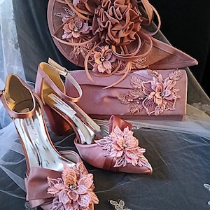 Puede incluir: Un conjunto de accesorios formales a juego en color rosa empolvado. El conjunto incluye un sombrero, un bolso de mano y un par de zapatos de tacón alto. Cada artículo está adornado con adornos florales y detalles intrincados, ideal para una ocasión especial.
