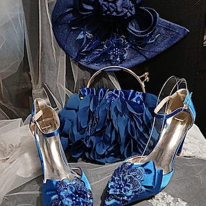 May include: A matching set of cobalt blue formal wear accessories. The set includes a hat, a handbag with a gold handle, and a pair of high-heeled shoes with floral embellishments. The shoes have a pointed toe and ankle straps. The accessories are on a white lace surface.