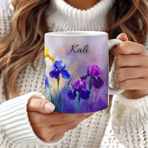 May include: White ceramic mug featuring a watercolor floral design with purple, blue, and yellow irises. The name "Kali" is written in elegant script. The mug is held by a person wearing a white sweater.