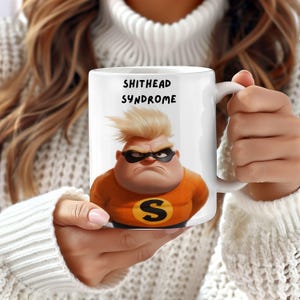 May include: White ceramic mug with a cartoon character wearing a superhero costume and the text "SHITHEAD SYNDROME".