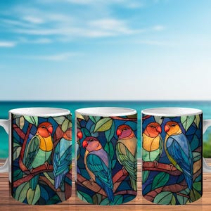 May include: White ceramic coffee mugs with a colorful stained-glass design featuring lovebirds perched on a branch with green leaves. The birds have orange, yellow, blue, and purple feathers. The background is a deep blue.