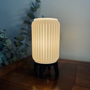 May include: A cream-colored, cylindrical lamp with vertical ribbed detailing. The lamp sits on a black, three-legged base. The lamp is illuminated, casting a warm glow. The lamp is on a dark wooden surface.