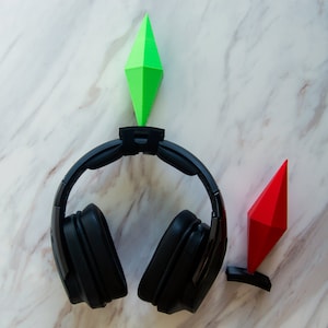 May include: Black over-ear headphones with a green and red diamond-shaped holder. The holders are designed to look like the plumbob from the Sims video game. The headphones are resting on a white and gray marble surface.