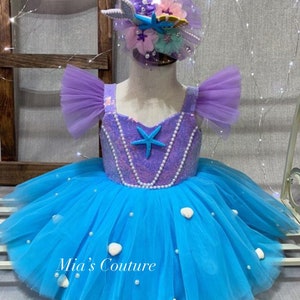 May include: A blue tulle dress with a purple bodice and white pearl accents. The bodice has a sequined mermaid tail design and a starfish. The dress is decorated with white seashells. The dress is for a child and has a matching headband with a starfish, seashells, and a mermaid tail.