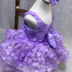 May include: A lavender tulle dress with 3D flower embellishments and a matching bow. The dress has a layered skirt and a sleeveless bodice.