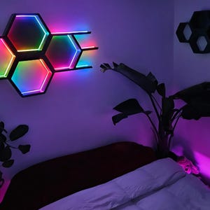 May include: A wall-mounted, black, hexagonal light fixture with color-changing LED lights. The fixture is composed of several connected honeycomb shapes, emitting a vibrant spectrum of colors. The room is decorated with plants and other hexagonal shelves.