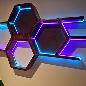 May include: A wooden, honeycomb-shaped wall shelf with built-in LED lighting. The shelves are stained a dark brown and feature blue and purple lights. The design is modern and geometric, suitable for displaying decor.