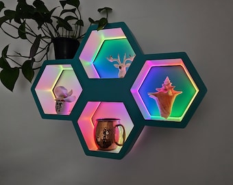May include: A teal, hexagon-shaped wall shelf with built-in LED lighting. The shelf displays a small deer figurine, a seashell, a succulent in a pot, and a copper mug. The LED lights cycle through various colors, creating a vibrant display.