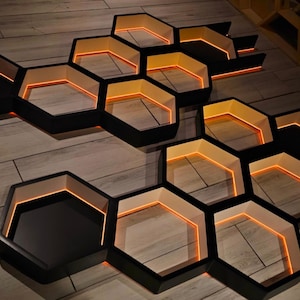 May include: A set of modular hexagonal wall shelves featuring a black exterior frame and a white interior lining. Each individual honeycomb-shaped unit is illuminated from within by a warm orange LED light strip, creating a glowing effect.