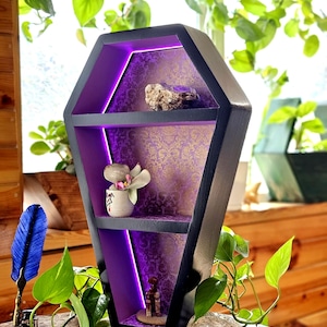 Pode incluir: Uma prateleira de parede preta em forma de caixão com um painel traseiro com padrão damasco roxo e dourado e iluminação LED roxa integrada. A prateleira tem dois níveis horizontais exibindo uma pequena pedra, um vaso de cerâmica e uma estatueta.