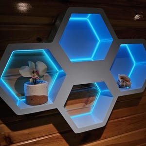 May include: A hexagonal shelf unit with blue LED lights, featuring a potted plant and a decorative seashell. The shelf is made of white wood and has a modern design.
