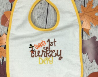 Puede incluir: Babero blanco para bebé con ribete amarillo. El babero tiene un pavo bordado con el texto "Owen's 1st Turkey Day".