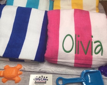 May include: Folded beach towels with bold stripes in blue, pink, and yellow. One pink towel is embroidered with the name "Olivia" in green. A blue sand shovel and orange turtle toy are visible.
