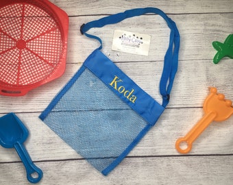 May include: Blue mesh beach bag with a blue strap and the name "Koda" embroidered in yellow. The bag is on a white wooden surface with a red plastic sieve, a blue plastic shovel, an orange plastic rake, and a green plastic starfish.