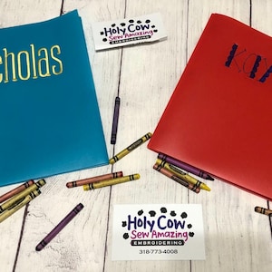 May include: Two personalized notebooks, one teal with "Nicholas" in gold lettering and one red with "Kaye" in blue lettering. Crayons are scattered around the notebooks. A business card for "Holy Cow Sew Amazing Embroidery" is also present.